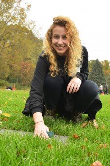Photo of a smiling female student who is leaning over an inground, gray grave marker amid a grassy field and using a green scour to remove its accumulated dirt.