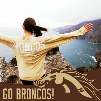A back portrait of a female student with Bronco T-shirt who is standing on a cliff facing the ocean.
