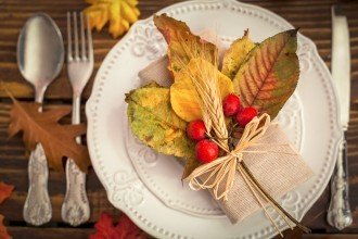 Photo of a placement setting on a table with a spoon, fork and knife and a plate with a festive fall leaves and berries decoration on it.