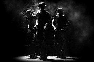 Black and white photo of three tap dancers wearing hats and simulating smoking on stage.
