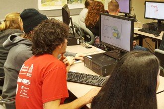 Photo of several groups of students gathered around computers as they work out geographic information science problems during an in-class GIS Challenge contest.