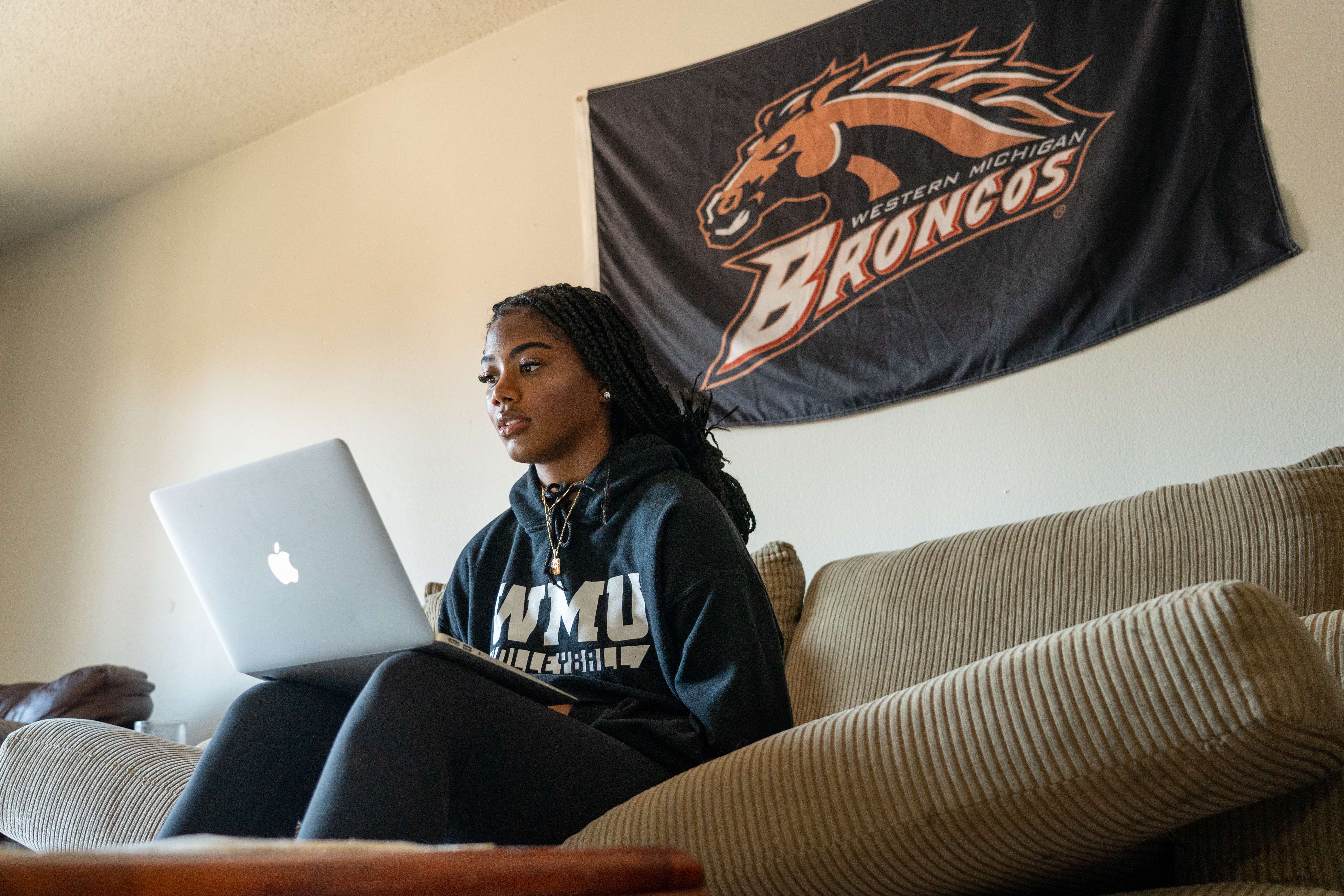 A student on her laptop sitting on the couch with a WMU Broncos sign hanging on the wall.