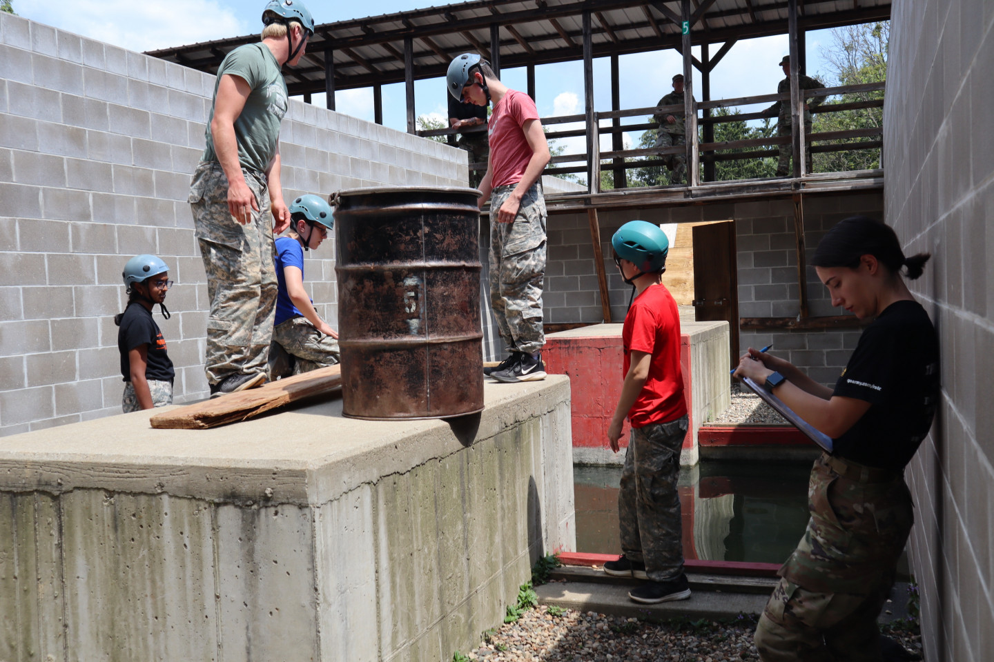 ROTC cadet Carolina Castanheira leans against a cement while campers stand on a cement slab with equipment for an obstacle.