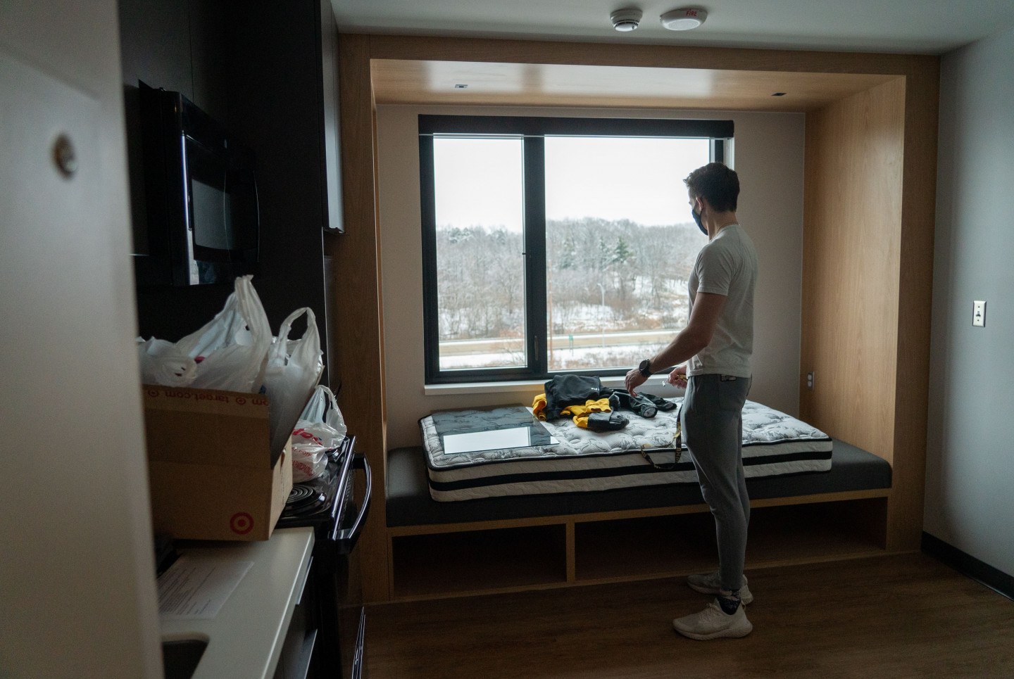 A student stands by a large window in his apartment.