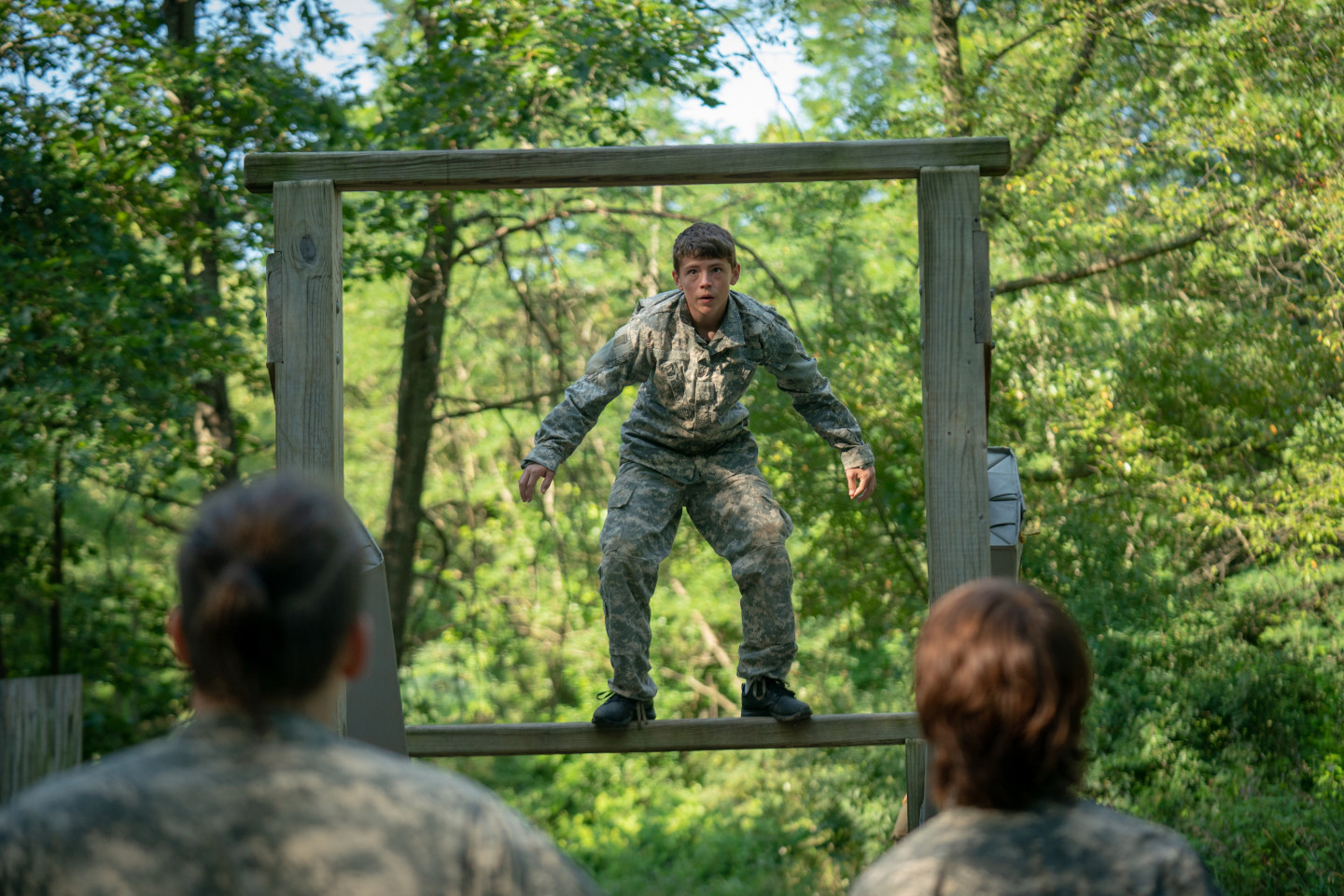 A teenager in Army fatigues stands in a wooden frame on an obstacle course.