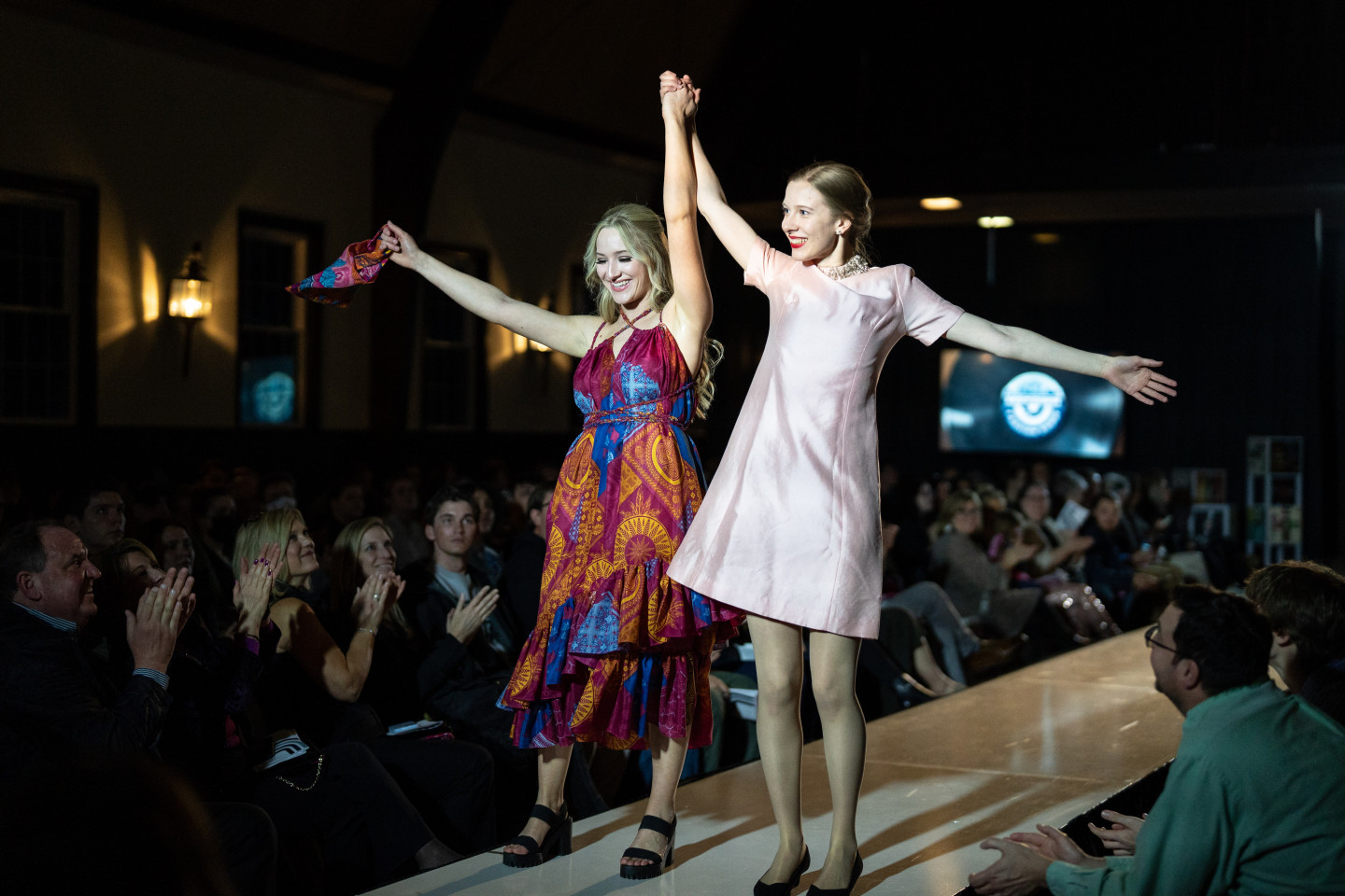 Two young women raise their hands on a fashion runway.