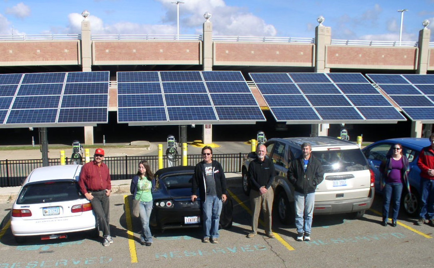 A group of people stand next to cars under a solar array.