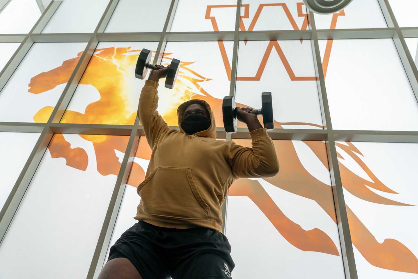 A man wearing a mask while lifting weights in front of the window of the Student Recreation Center.