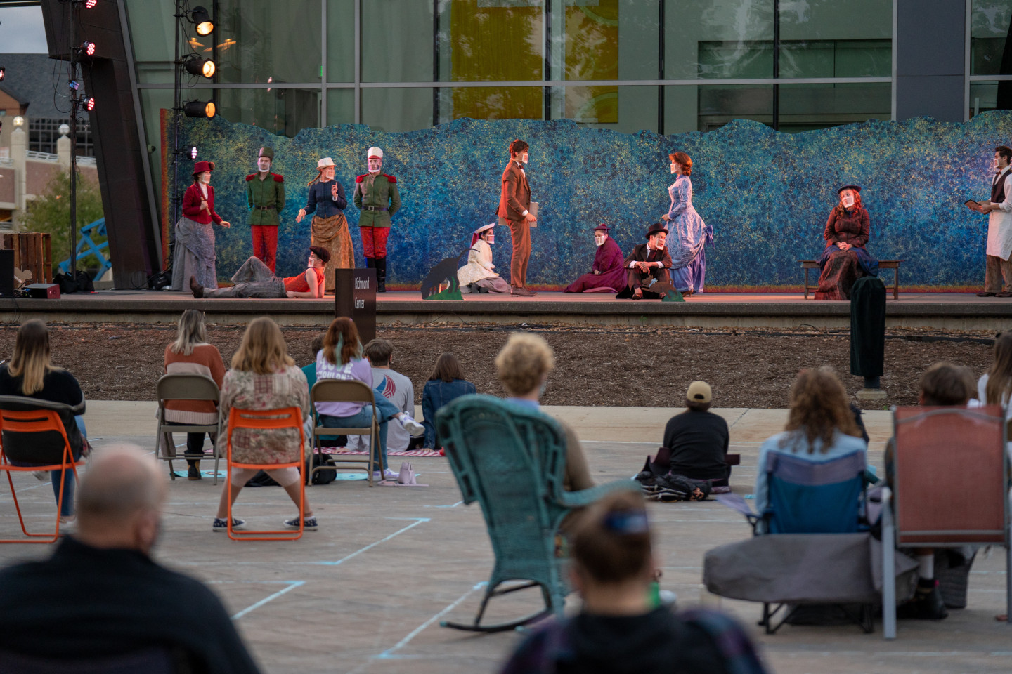 A socially distanced crowd watches actors wearing masks perform on an outdoor stage.