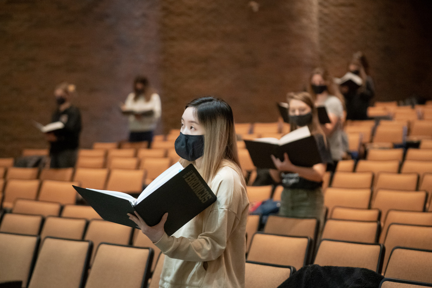 Students wearing masks sing while holding sheet music in a lecture hall.