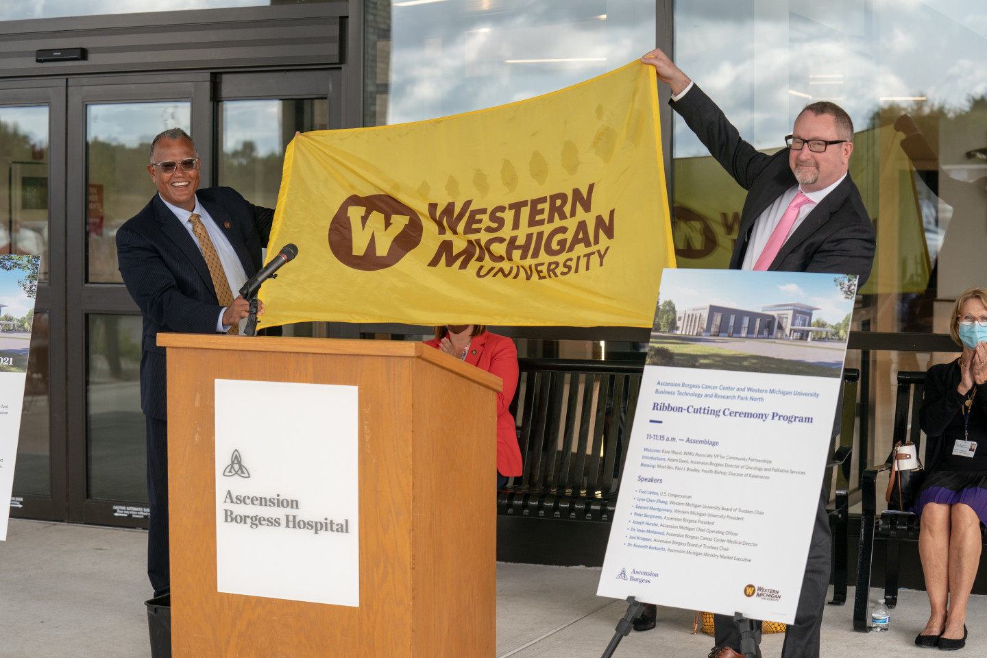 President Montgomery holds up a Western Michigan University flag with President Peter Bergmann of Ascension Borgess in front of the podium of the Ascension Borgess Cancer Center.