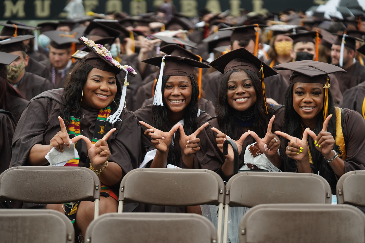 Four students in caps and gowns hold up their fingers in Ws.