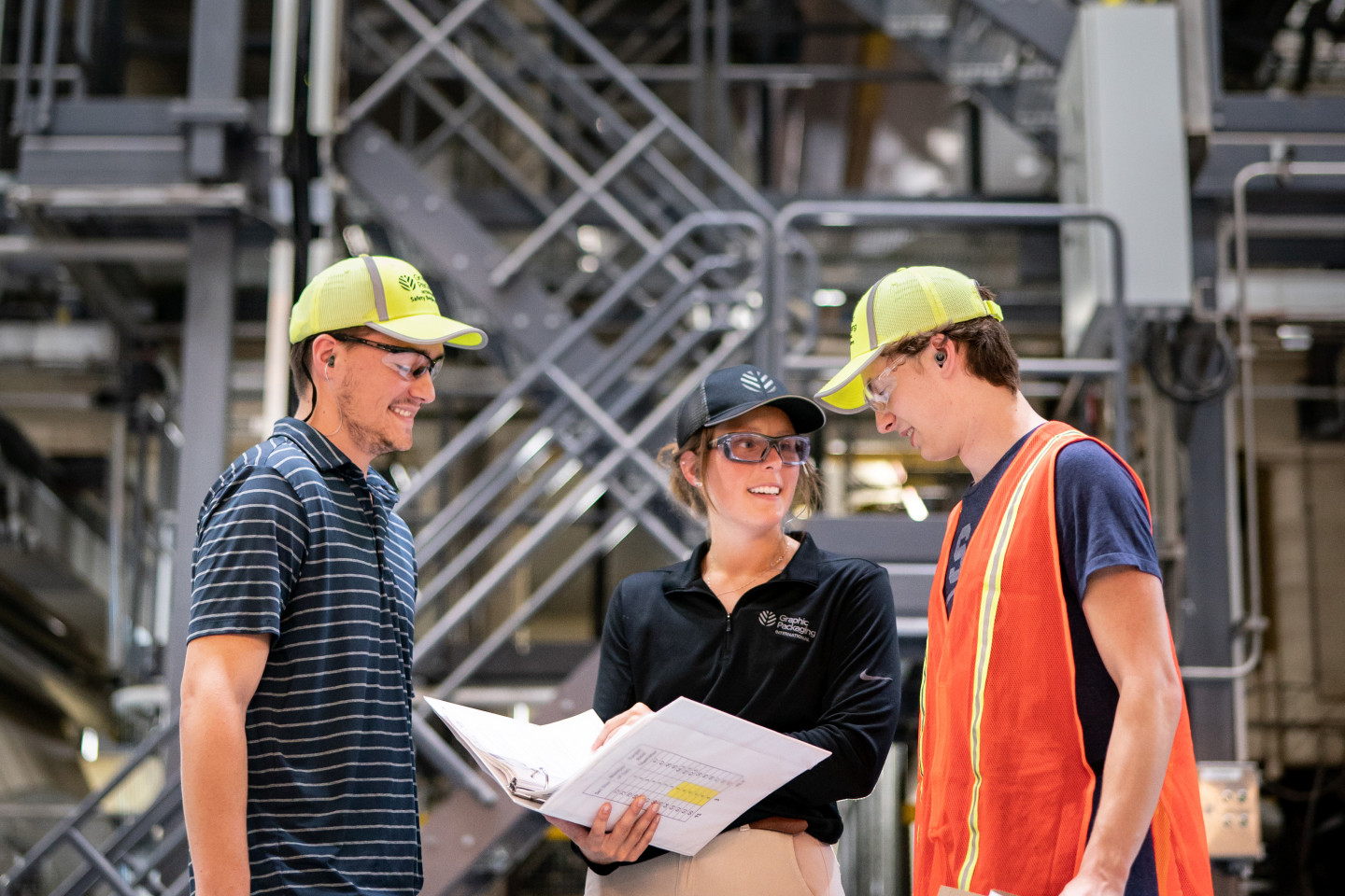 Hannah Kalleward holds a binder and talks with two other students wearing hard hats in an industrial setting.