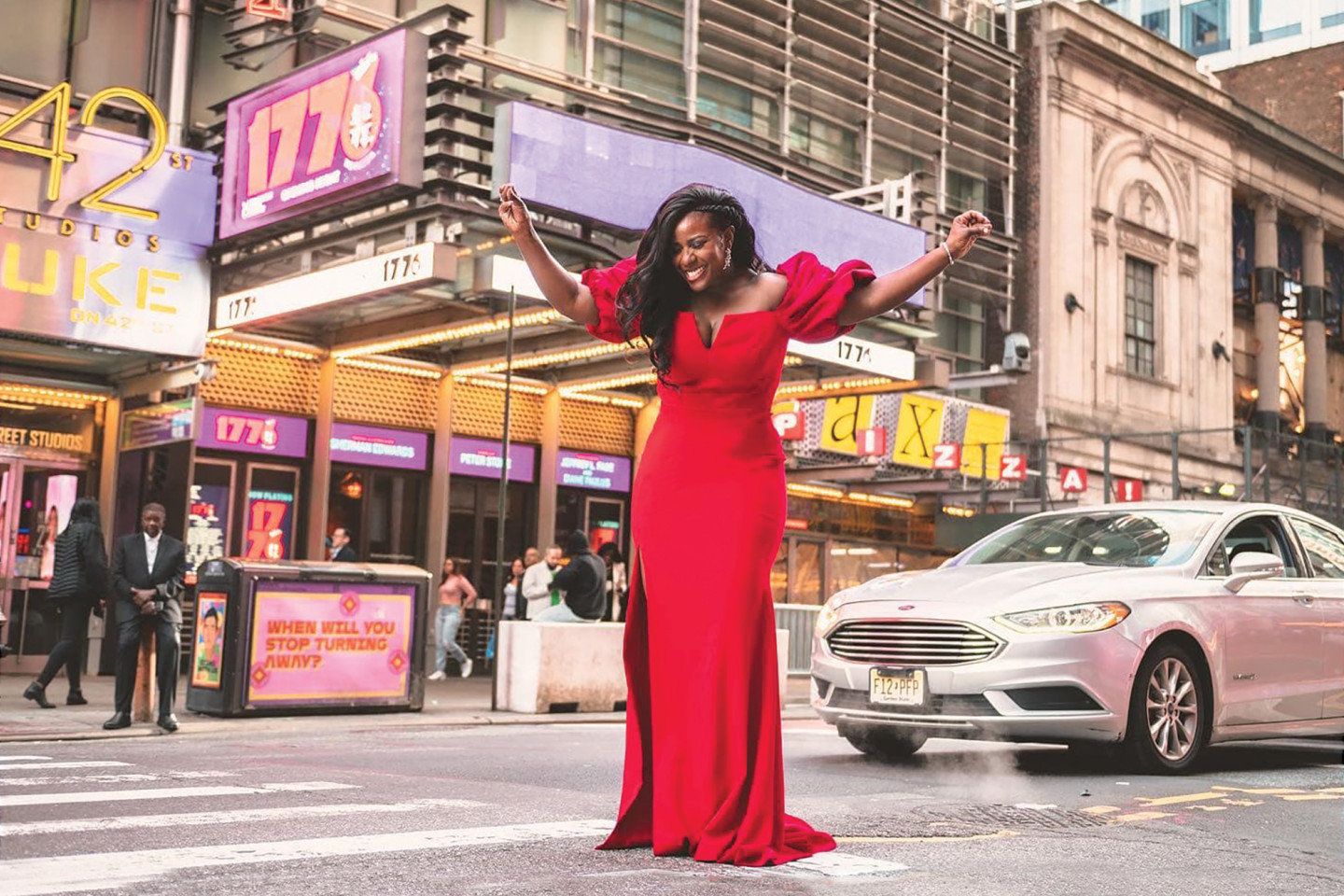 Crystal Lucas-Perry stands in a crosswalk in front of a Broadway theatre wearing a red dress. Photo courtesy: Valerie Terranova. 