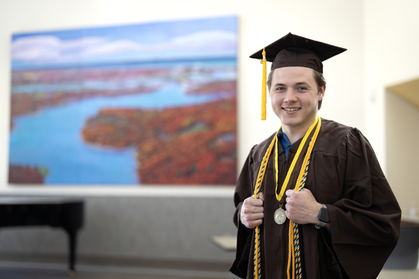 A portrait of Quinn Heiser in his graduation cap and gown standing in front of a scenic painting.