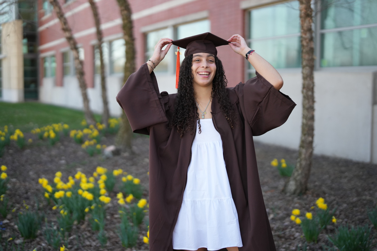 Zahi Sanchez poses outside in a graduation cap and gown with yellow flowers in the background.