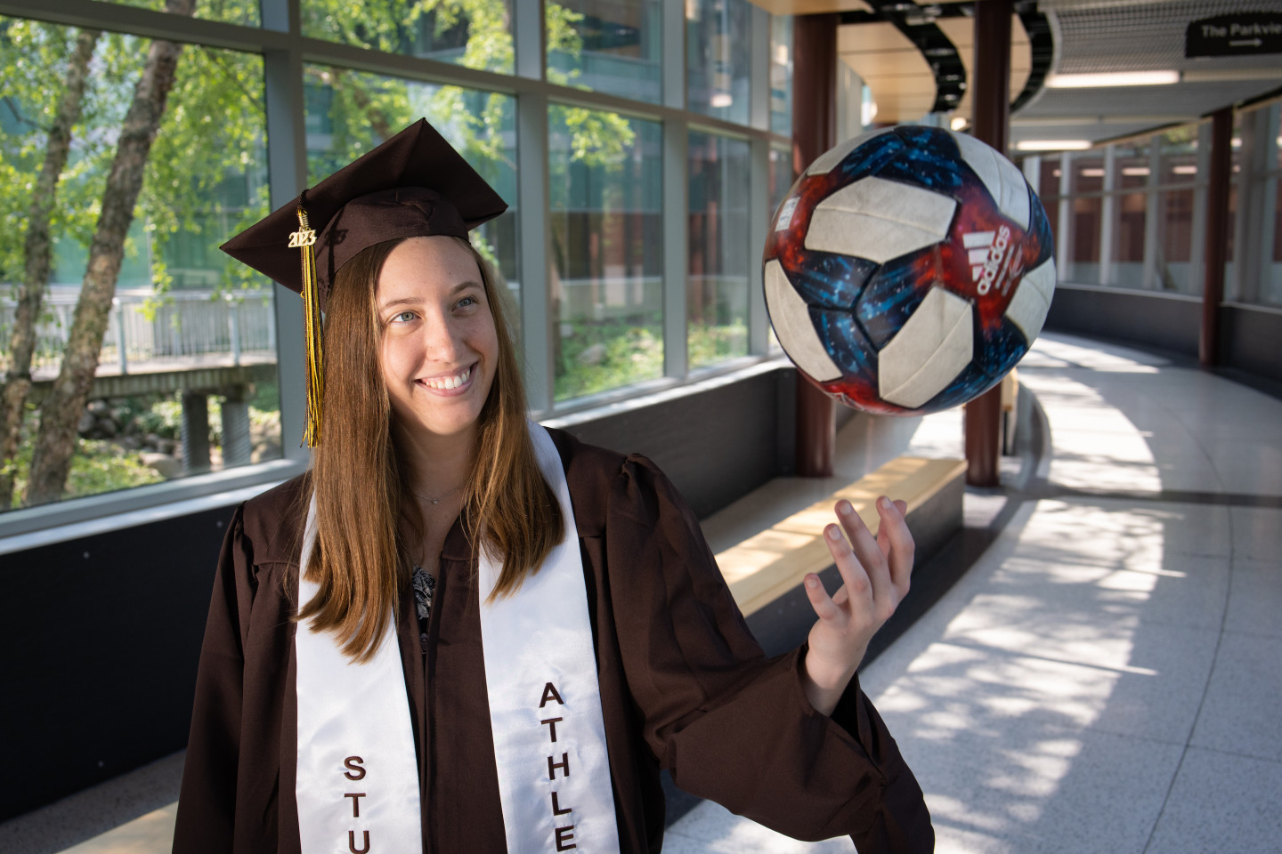 Hannah Sargent tosses a soccer ball in the air while wearing her graduation cap and gown.