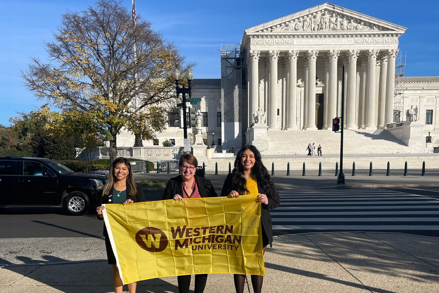 Three people hold a gold-colored Western Michigan University flag in front of the U.S. Supreme Court.