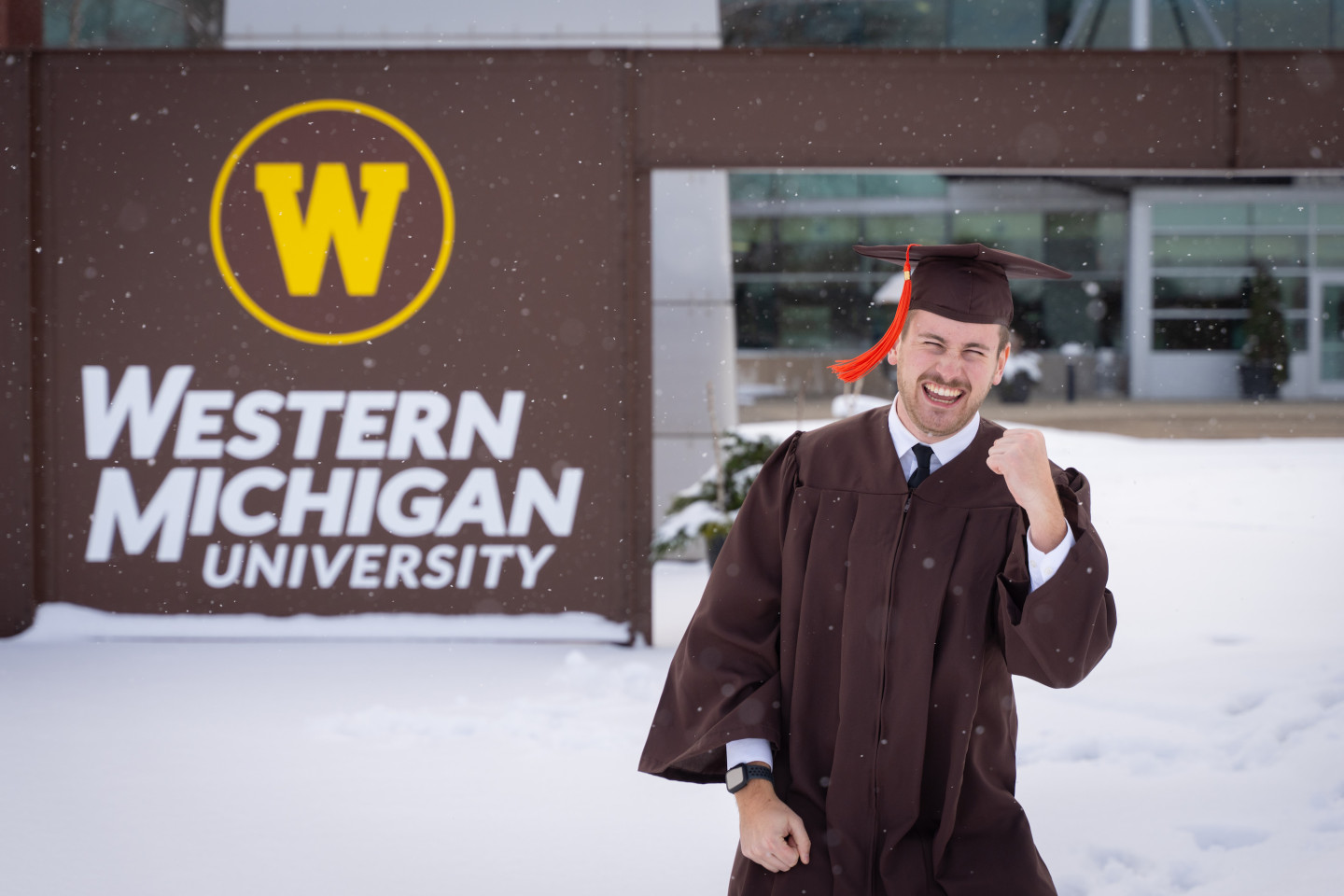 Joh Goheen raises a fist in celebration while standing outside in his graduation regalia as the snow falls.