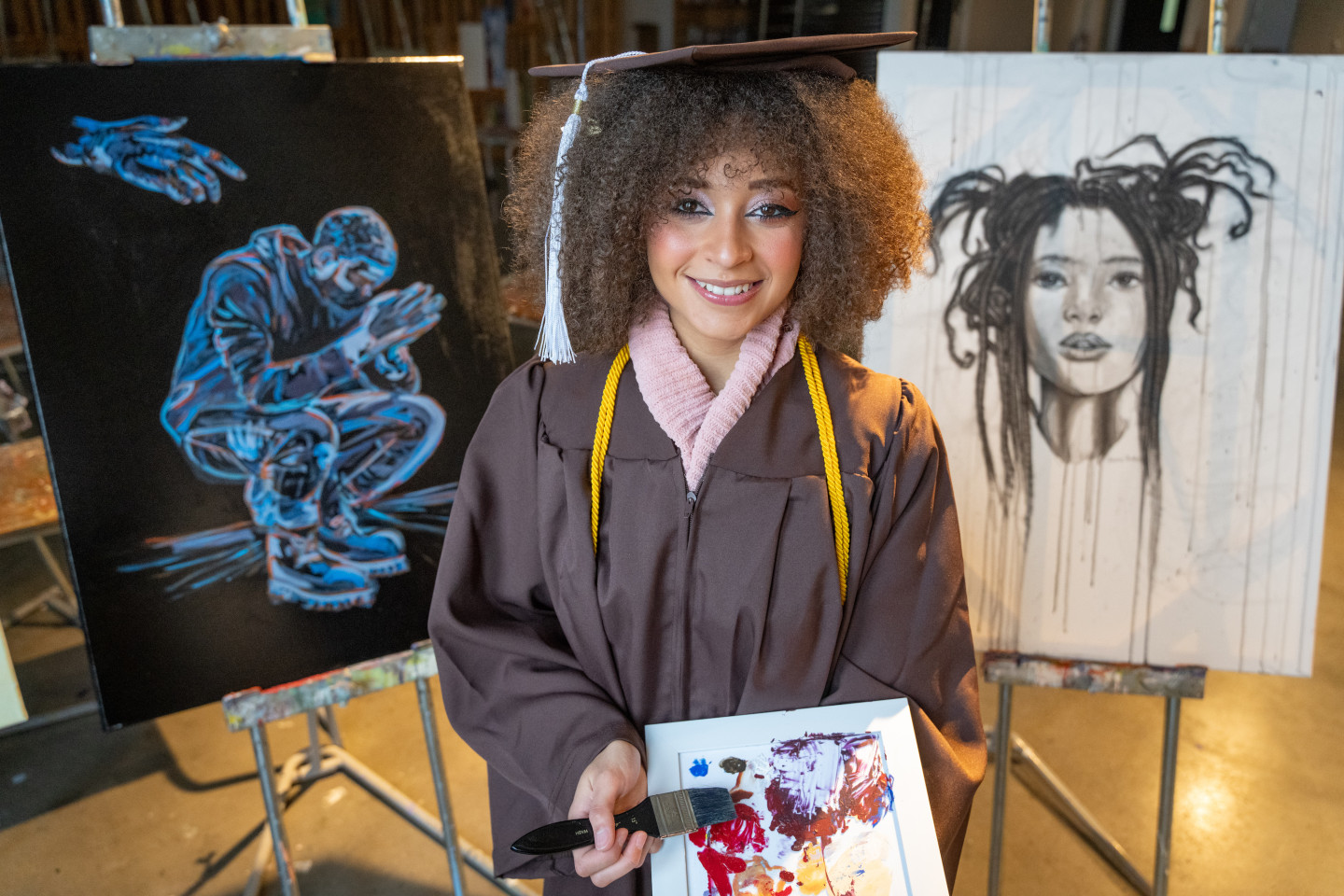 A portrait of Sharmane Flanders in her graduation regalia standing in front of two paintings and holding a colorful canvas and paint brush.