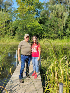 Two people stand on a dock in a luch, green vegetative setting.