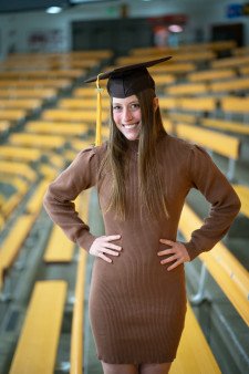 Grace Filpi poses with her hands on her hips while wearing a graduation cap and brown dress. There are yellow bleachers in the background.