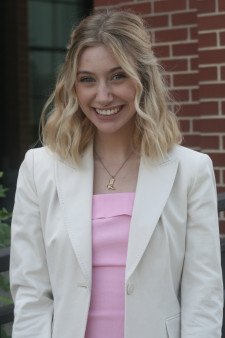 Photo of Liv Hale standing outside in front of a brick wall, wearing a white blazer and pink blouse