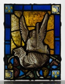 A fragment of a stained glass window in tones of blue and gold depicting an angel robed in white playing a portatif organ. 