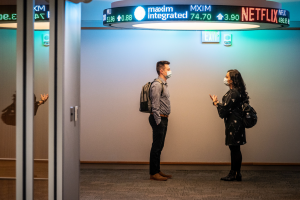 Two masked students standing in hallway under stock ticker