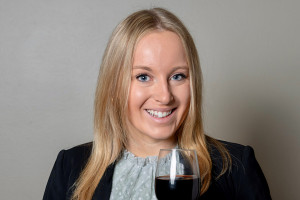 Megan Barr is wearing a black blazer, holding a glass of red wine.