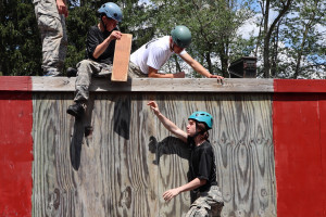 Campers sit atop a wall while handing a plank to a camper on the ground below.