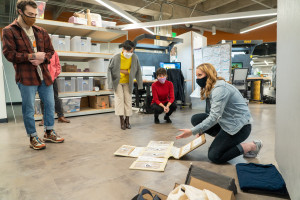 A student kneels down to present designs on cardboard on the ground.