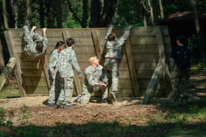 Teenagers in Army fatigues help each other scale a wall on an obstacle course.
