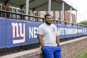 Brandon Harlin leans against a fence with a banner that reads, "NY New York Giants Training Camp."