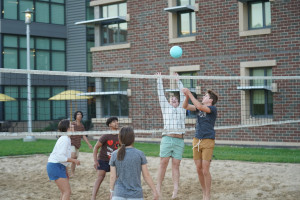 Students play volleyball on a sand court.