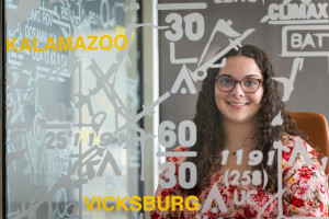 Emily Hartzell sits behind a pane of glass with graphics on it and the cities of Kalamazoo and Vicksburg.