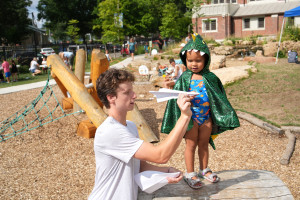 Gabe Bresnahan shows a young girl wearing a dinosaur cape how to throw a paper airplane.