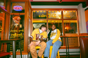 Two students sit on stools inside a pizza shop.
