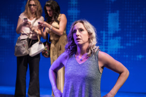 Kate Thomsen stands with her arms on her hips while two women stand in the background looking at their mobile phones.