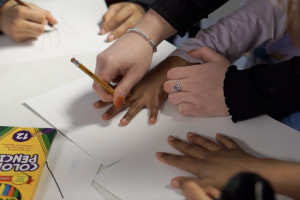 An adult hand traces a child's hand on a piece of paper with a pencil.