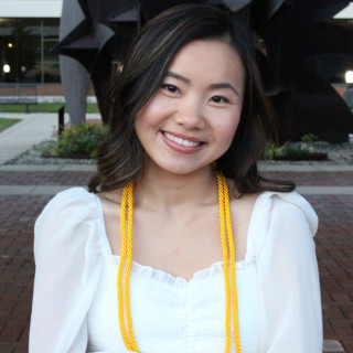 Julie Schwark is wearing a white dress, with gold honor cords, outside of the Haworth College of Business.