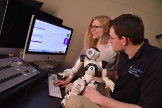 Communication students holding a robot while looking at its diagnostics on a computer screen