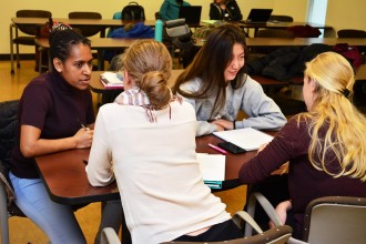 four female students from diverse ethnicity are sitting in a classroom and talking together. 