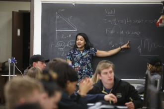 An instructor points to physics equations on a chalkboard.