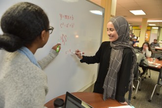 Two women write mathematical equations on a white board.