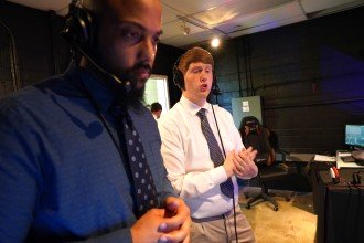 Two men dressed in suits wear headsets and provide commentary into microphones.