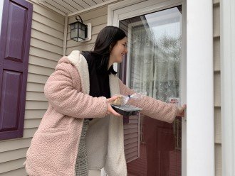 A young woman rings a doorbell as she holds a hot meal she is delivering to a senior.