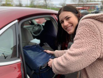 A young woman smiles as she loads a cooler of food into the back seat of a car.