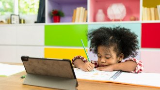 A child writes on a piece of paper while watching a tablet.
