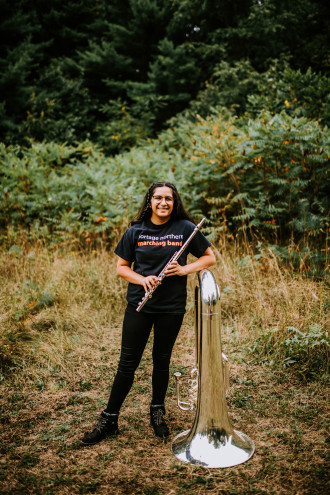Keshavi Davé stands in nature, holding her flute and leaning on her tuba.